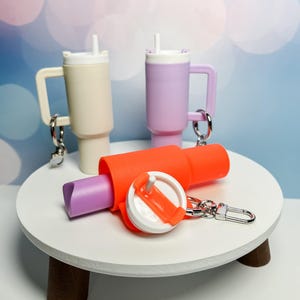 May include: Three miniature plastic cups with lids and straws, one beige, one purple, and one orange, are attached to a keychain. The keychain is on a white table with a brown base.