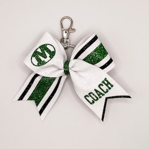 May include: White cheer bow with green and black stripes and glitter. The bow has a green "M" in a circle on one side and the word "COACH" on the other side. The bow is attached to a silver key chain.