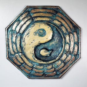 May include: Octagonal wooden wall decor with a raised, carved yin yang symbol in gold and teal green. The yin yang is surrounded by concentric circles of raised, carved lines in gold and teal green.