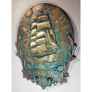 May include: A gold and teal colored metal wall plaque with a raised image of a ship with sails and the text "HOLD FAST".