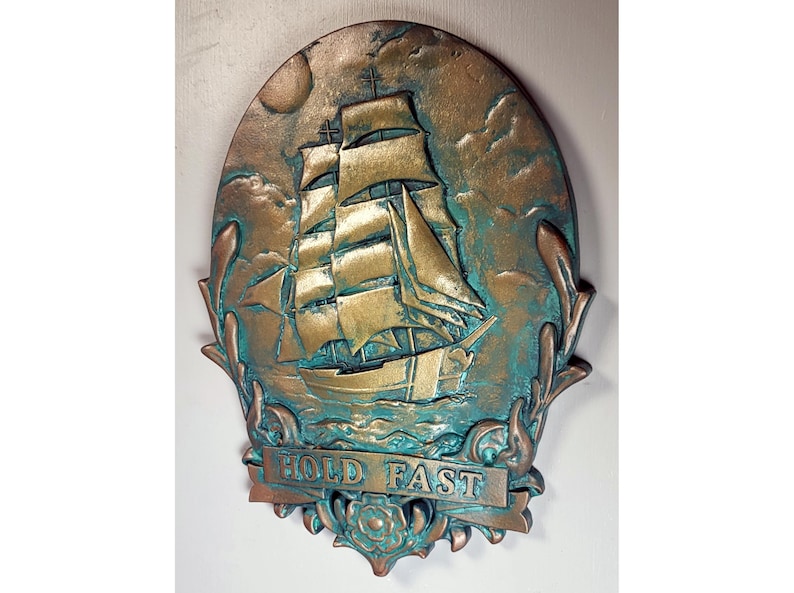 May include: A gold and teal colored metal wall plaque with a raised image of a ship with sails and the text "HOLD FAST".