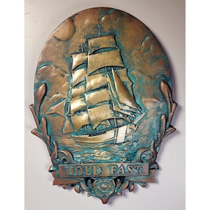 May include: A circular, bronze-colored wall plaque with a raised image of a ship with sails. The ship is surrounded by a decorative border of leaves and a banner that reads "HOLD FAST".