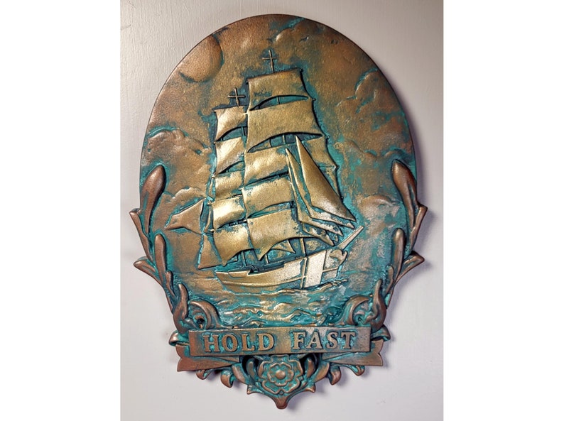 May include: A circular, bronze-colored wall plaque with a raised image of a ship with sails. The ship is surrounded by a decorative border of leaves and a banner that reads "HOLD FAST".