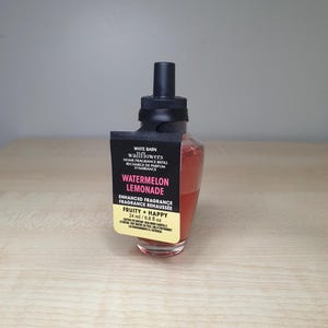 May include: A clear glass bottle with a black cap and a pink liquid inside. The bottle has a label that says "White Barn Wallflowers Home Fragrance Refill Watermelon Lemonade Enhanced Fragrance Fragrance Rehaussée Fruity + Happy 24 ml / 0.8 fl oz".