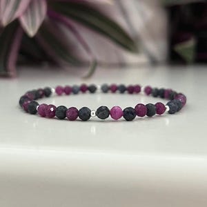 May include: A bracelet made of alternating dark blue and magenta gemstones. The beads are strung on a thin, silver-coloured wire. The background shows a blurred plant with green and purple leaves.