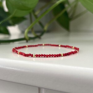 May include: A delicate red beaded bracelet with gold-coloured accents. The bracelet features small, faceted red beads interspersed with gold-coloured beads. The jewellery is displayed on a white surface, with green foliage in the background.