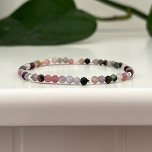 May include: A delicate bracelet featuring small, colourful gemstone beads. The beads are in shades of pink, purple, green, and black. The bracelet is strung on a silver chain.