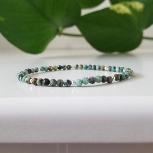 May include: A delicate beaded bracelet with small, round beads in teal, brown, and black. The beads are strung on a thin, silver-coloured wire, creating a minimalist design. The bracelet is displayed on a white surface. A stylish accessory.