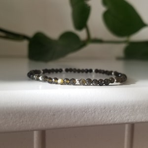 May include: A delicate bracelet made of small, dark grey and gold beads. The bracelet is on a white surface.