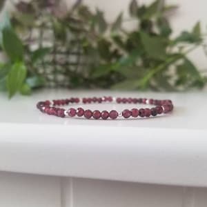 Peut inclure: Un bracelet délicat composé de petites perles de grenat rouge foncé et d'entretoises argentées. Le bracelet est posé sur une surface blanche.