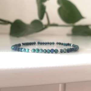May include: A beaded bracelet featuring faceted, round beads in shades of blue and green. The bracelet is strung on a thin, elastic cord with small silver-colored spacer beads. The bracelet is approximately 7 inches in length.