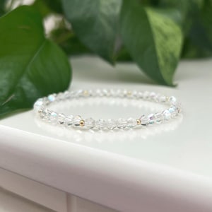 May include: A clear crystal bracelet with small, faceted beads and gold-coloured accents. The bracelet is resting on a white surface, with green foliage in the background. The bracelet appears to be approximately 18 cm in circumference.