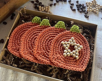 May include: A set of four orange crochet pumpkin coasters with green stems and white bead accents, presented in a cardboard box with shredded paper. The coasters are handmade and suitable for fall decor.