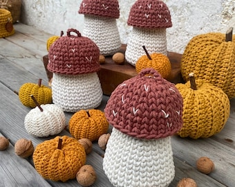 May include: A collection of crocheted mushroom ornaments in various shades of brown and white. The mushrooms are arranged on a wooden surface with other crocheted items, including a basket and a small pumpkin.
