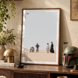Op de afbeelding: Een ingelijste kunstprint met een scène uit Star Wars met R2-D2, C-3PO, Prinses Leia en Luke Skywalker staand in het zanderige woestijnlandschap van Tatooine onder een heldere lucht, gepresenteerd in een eenvoudige houten lijst.