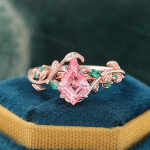 May include: Rose gold ring featuring a kite-shaped pink gemstone as the centerpiece. The ring is adorned with leaf and vine detailing, accented by small green gemstones. The ring is set on a blue velvet surface.