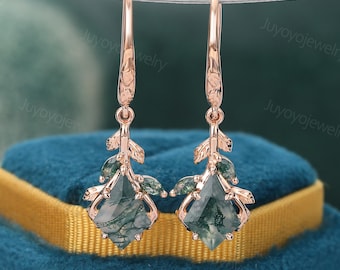 May include: Rose gold drop earrings with green moss agate gemstones. The earrings feature a leaf design with a geometric cut gemstone at the centre. The earrings are suspended from a hook.