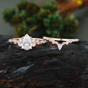 May include: A rose gold engagement ring set featuring a pear-cut center stone surrounded by smaller round stones on a leaf-textured band, accompanied by a matching V-shaped wedding band with a small pear-cut stone and leaf-like detailing.