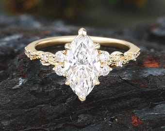 May include: A gold ring featuring a large marquise-cut center stone flanked by three smaller round stones on each side. The band is crafted with a textured, organic leaf-like design that extends along the sides, set with tiny accent stones.