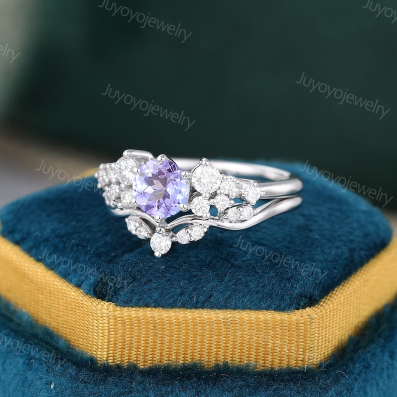 May include: A delicate silver ring set with a round, light purple gemstone surrounded by smaller clear gemstones. The ring is set on a blue velvet background.