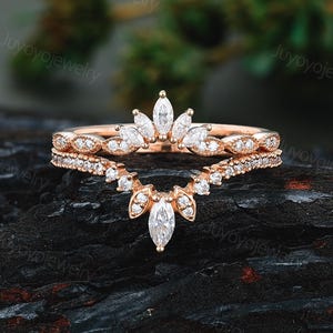 May include: A rose gold ring set consisting of two stacked bands. The top band features marquise-cut stones in a crown arrangement, while the bottom band is a V-shaped chevron style adorned with small round stones and a central marquise-cut stone.