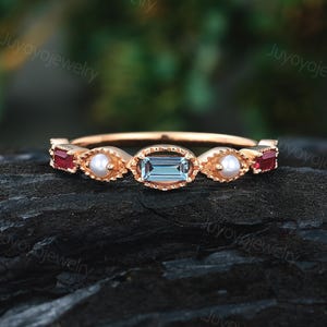 May include: A gold ring featuring an aquamarine baguette center stone, flanked by two red gemstones and two pearls. The ring has a delicate band and is set on a dark, textured surface. The ring is a piece of jewelry.