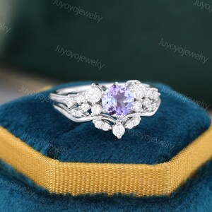 May include: A silver ring set with a round, purple amethyst gemstone surrounded by smaller, clear diamonds. The ring is on a blue velvet box.