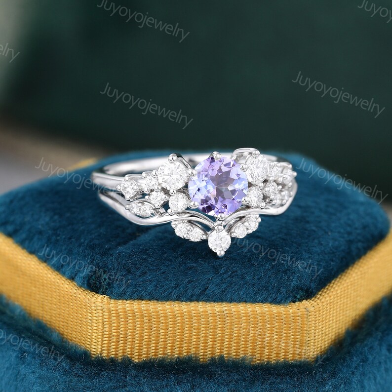 May include: A silver ring set with a round, purple amethyst gemstone surrounded by smaller, clear diamonds. The ring is on a blue velvet box.