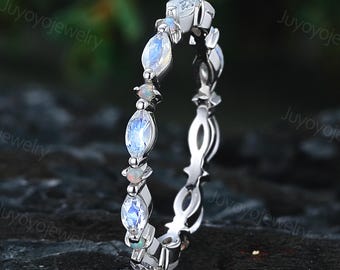 May include: A silver ring featuring alternating oval and round gemstones. The ring has a delicate design with a series of oval-shaped, light blue gemstones and smaller, round, white gemstones. The ring is set against a dark background.