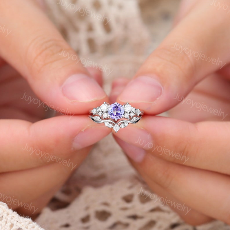May include: A delicate silver ring set with a purple gemstone surrounded by smaller clear gemstones. The ring is designed with a delicate floral motif.
