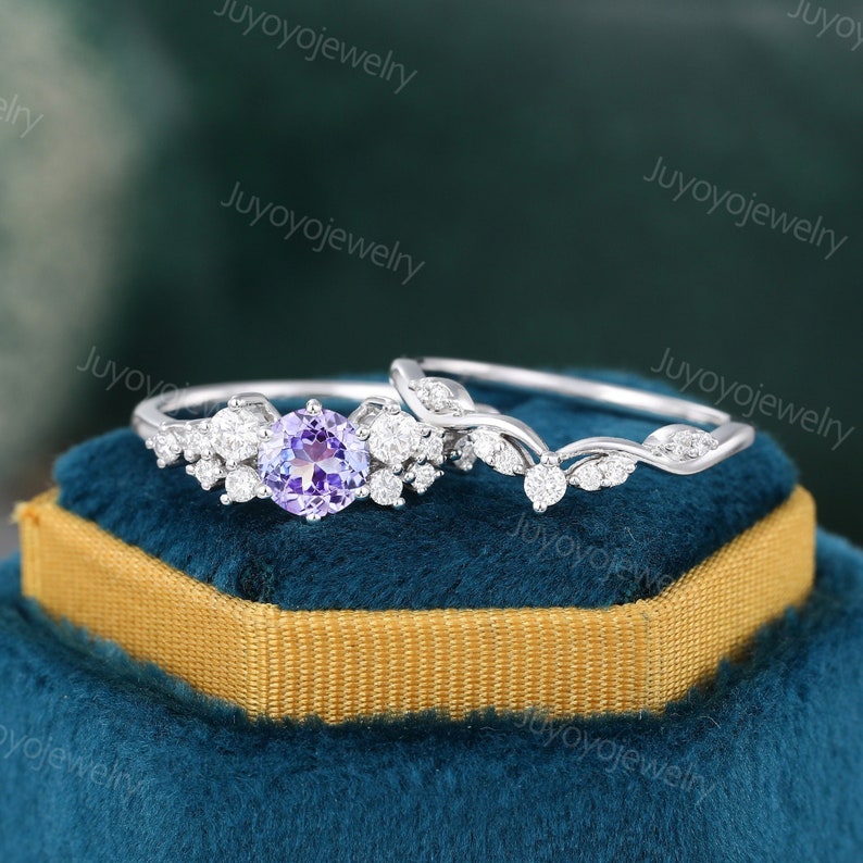 May include: A delicate silver engagement ring set with a purple amethyst center stone and a matching wedding band with a vine design. The rings are displayed on a blue velvet box with a gold trim.