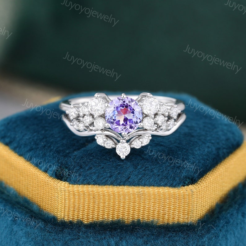 May include: A silver ring set with a purple gemstone surrounded by smaller clear gemstones. The ring is on a blue velvet box.