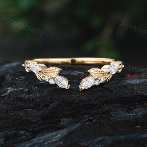 Peut inclure: Une bague dorée avec un design à tige fendue. La bague présente des pierres précieuses blanches de taille marquise et ronde serties dans des grappes en forme de feuilles de chaque côté de la fente. La bague est présentée sur une surface sombre et texturée.