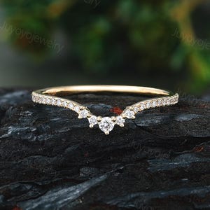 Puede incluir: Un anillo dorado con un diseño curvo, engastado con pequeños diamantes brillantes. El anillo presenta un grupo central de tres diamantes más grandes, creando una apariencia delicada y elegante. El anillo se muestra sobre una superficie oscura y texturizada.