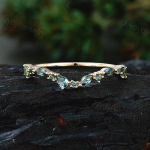 May include: A gold band ring featuring a curved, V-shaped design set with multiple small, pear-shaped and round green gemstones. The stones are arranged in a cluster along the top arc of the band, showcasing a polished gold metal finish.