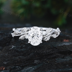 May include: A silver engagement ring with an oval-cut diamond center stone. The band features a leaf and vine design with small accent stones. The ring is set against a dark, textured background, highlighting the sparkle of the diamond.