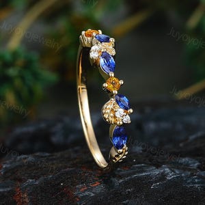 May include: A gold ring featuring a series of marquise-cut blue sapphires, round yellow sapphires, and small clear white stones arranged in a curved pattern along the band, accented with delicate gold leaf-shaped metal details.
