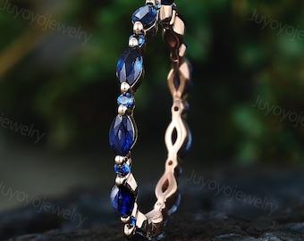 May include: A rose gold ring featuring alternating marquise and round-cut blue gemstones. The gemstones are set in a continuous pattern around the band. The ring is displayed against a dark, textured surface with a blurred green background.