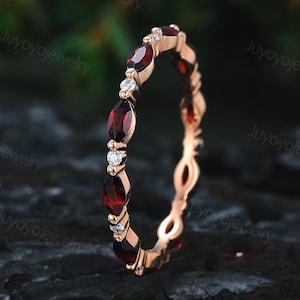 Op de afbeelding: Een roségouden ring met afwisselend marquise-geslepen rode edelstenen en kleine, ronde, heldere edelstenen. De ring heeft een delicaat, verweven bandontwerp. De ring wordt gepresenteerd op een donker, getextureerd oppervlak.