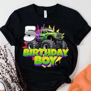 May include: Black t-shirt featuring a green and purple monster truck design with the number 5 and the words "BIRTHDAY BOY" in a colorful font. The design includes splashes of color.