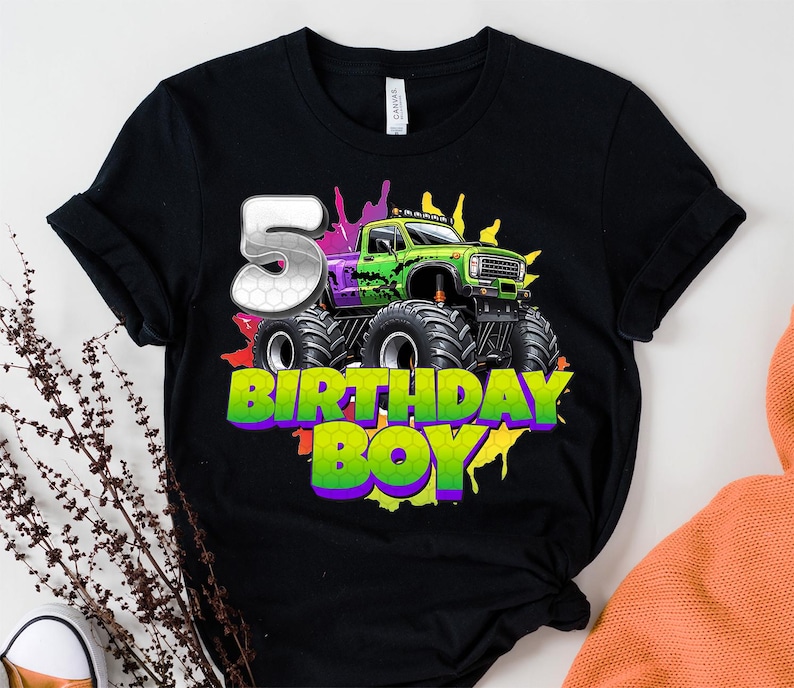 May include: Black t-shirt featuring a green and purple monster truck design with the number 5 and the words "BIRTHDAY BOY" in a colorful font. The design includes splashes of color.
