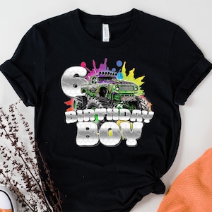 May include: Black t-shirt featuring a colorful monster truck design with the text "6 BIRTHDAY BOY." The graphic includes a green and purple monster truck with a splash of colors in the background. The number 6 is in a silver honeycomb pattern.