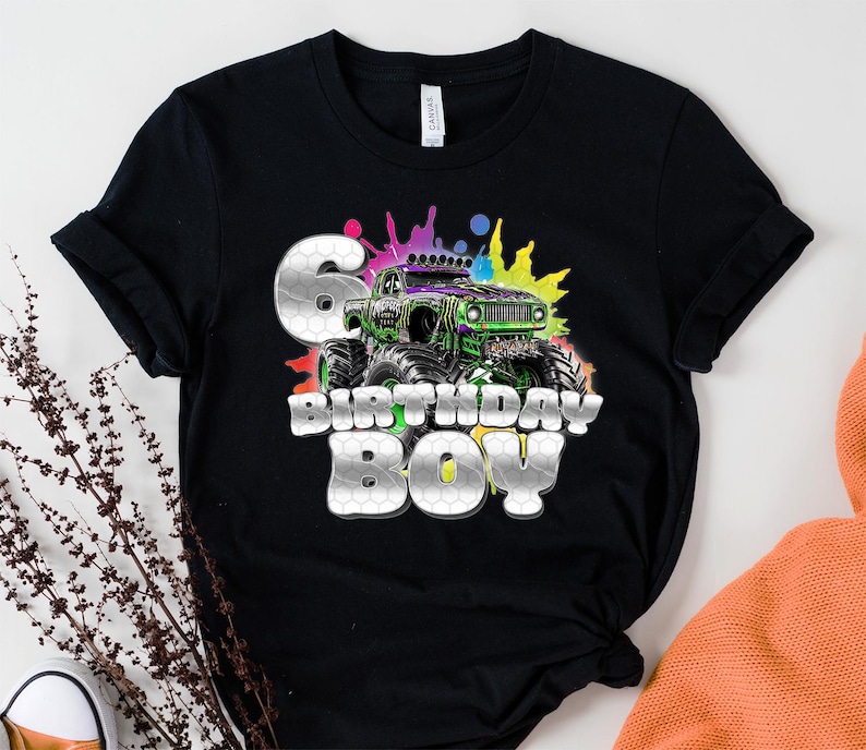 May include: Black t-shirt featuring a colorful monster truck design with the text "6 BIRTHDAY BOY." The graphic includes a green and purple monster truck with a splash of colors in the background. The number 6 is in a silver honeycomb pattern.