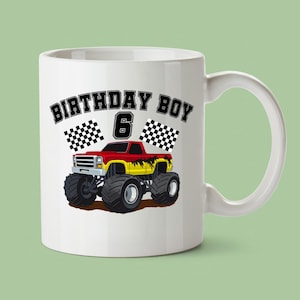 May include: White ceramic mug featuring a red, yellow, and black monster truck design. The mug has the text "BIRTHDAY BOY 6" with checkered flags. The mug has a white handle and is set against a light green background.