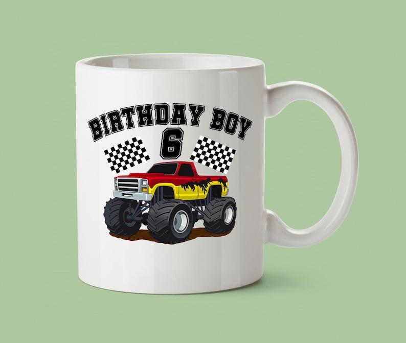 May include: White ceramic mug featuring a red, yellow, and black monster truck design. The mug has the text "BIRTHDAY BOY 6" with checkered flags. The mug has a white handle and is set against a light green background.