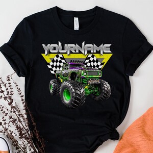 May include: Black t-shirt featuring a monster truck graphic with the text "YOURNAME" in silver and yellow. The monster truck is green and purple with large black tires and green rims. Checkered flags are on either side of the text.