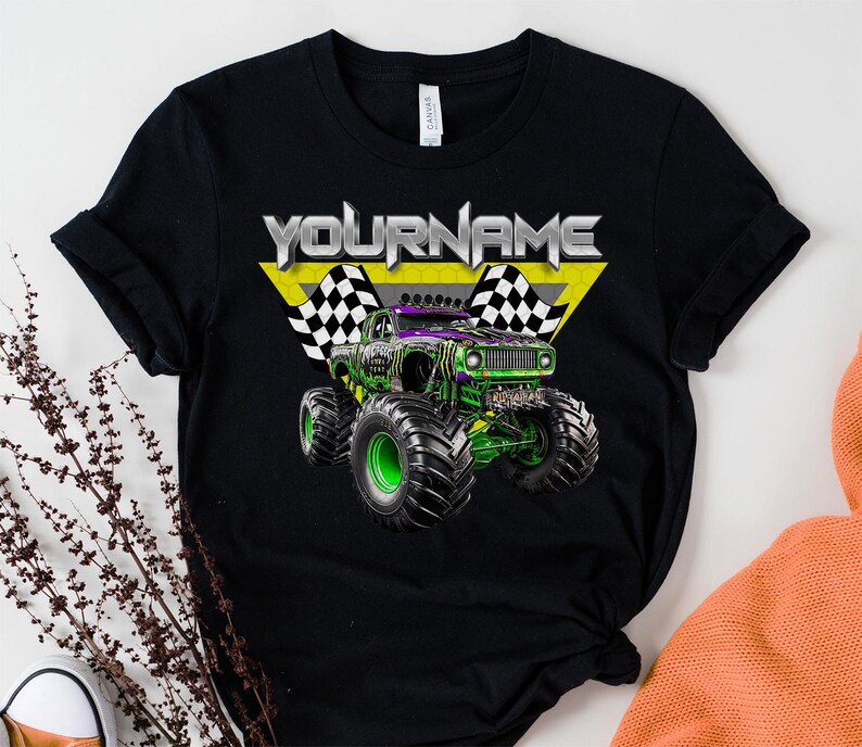 May include: Black t-shirt featuring a monster truck graphic with the text "YOURNAME" in silver and yellow. The monster truck is green and purple with large black tires and green rims. Checkered flags are on either side of the text.