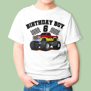 May include: White t-shirt featuring a colorful monster truck graphic. The design includes the text "BIRTHDAY BOY 6" with checkered flags. The truck is red and yellow, with large black tires.