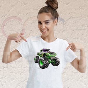 May include: White t-shirt with a green and purple monster truck graphic. The truck has large tires and a black grill.