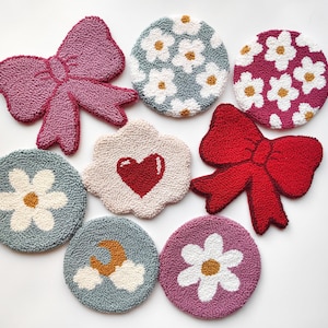 Può includere: Otto sottobicchieri rotondi fatti a mano con diversi disegni. I disegni includono un fiocco rosa, un fiocco rosso, un cuore bianco su sfondo crema, un fiore bianco su sfondo blu, un fiore bianco su sfondo rosa, un fiore bianco su sfondo blu, una luna bianca e nuvole su sfondo blu, e un fiore bianco su sfondo rosa.